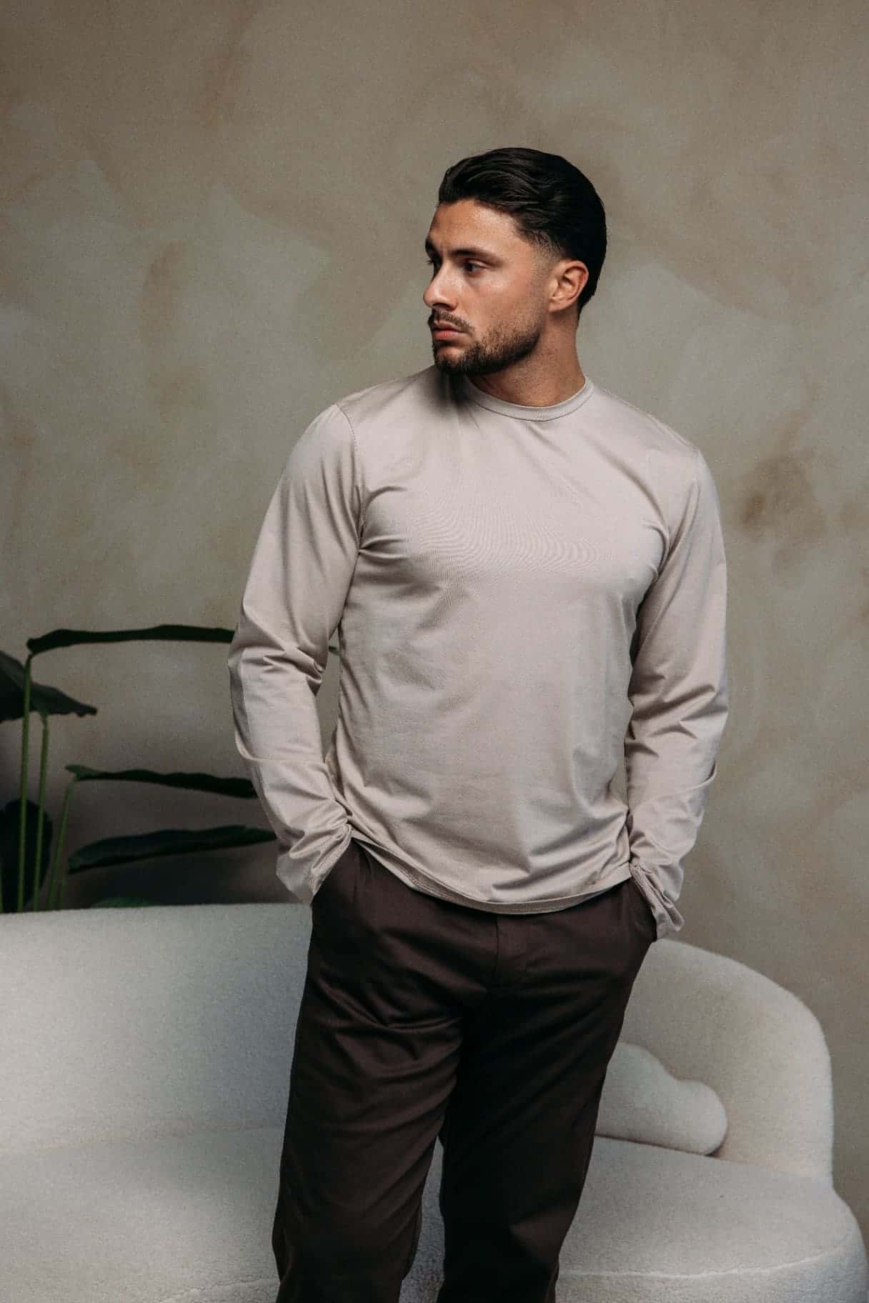 Model in a beige longsleeve and dark brown trousers in front of a bouclé sofa