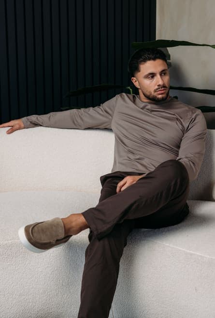 Model in a taupe long sleeve and brown trousers on a white sofa
